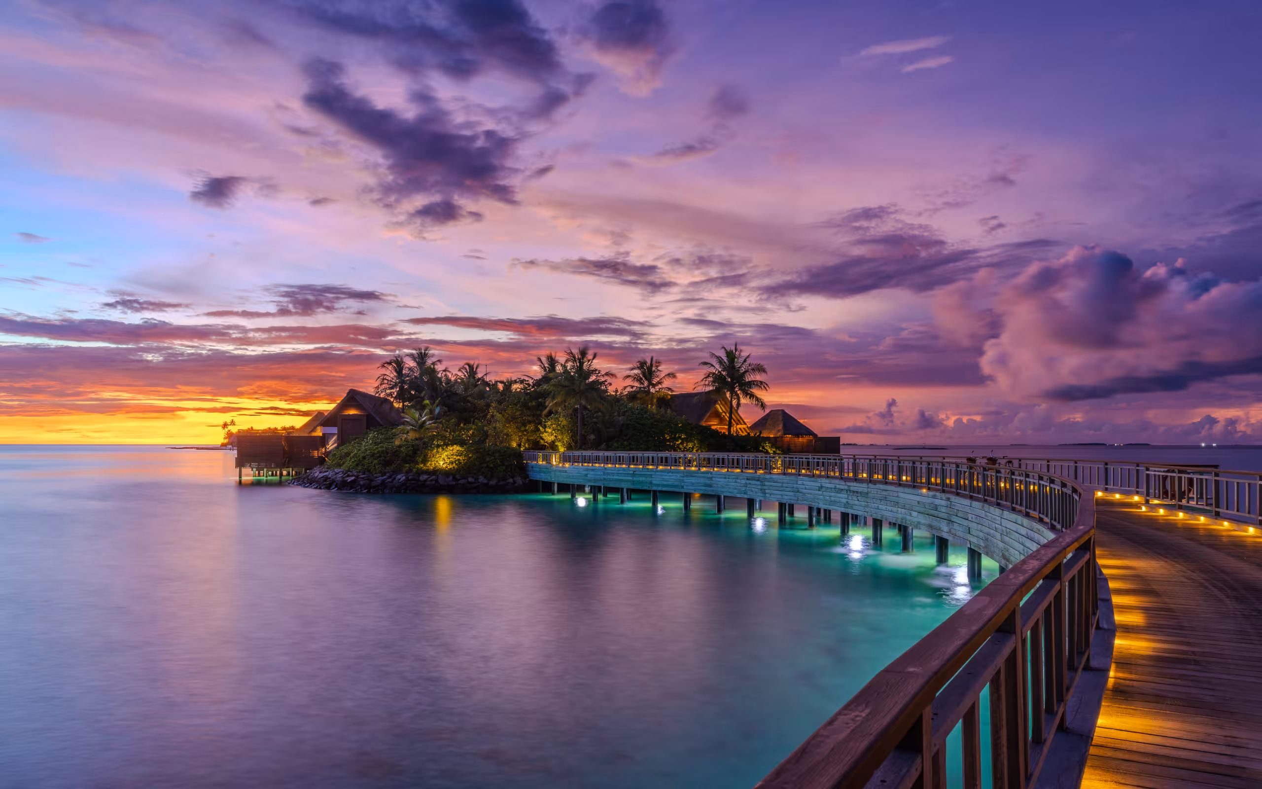 A small private island connected to the mainland by a snaking wooden bridge. The bridge is lit with coloured lights under the waters edge.